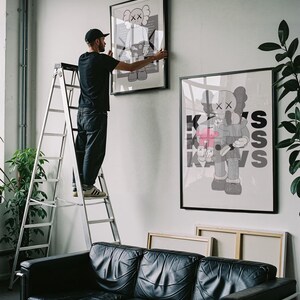 Puede incluir: Una persona en una escalera cuelga una obra de arte enmarcada en una pared blanca. La obra presenta una figura de dibujos animados con los ojos tachados. Debajo hay un sof&aacute; de cuero negro. Tambi&eacute;n se ven obras de arte enmarcadas adicionales y plantas.
