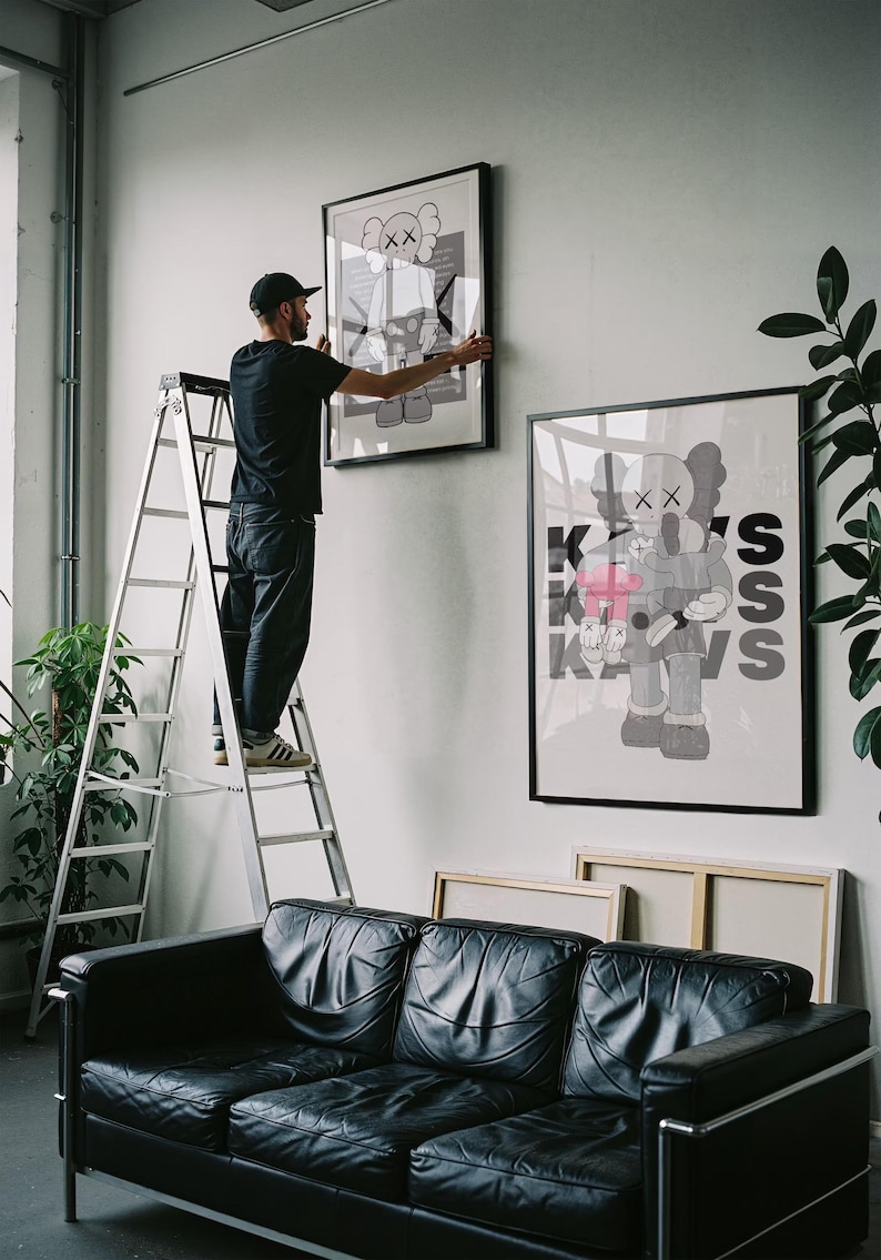 Puede incluir: Una persona en una escalera cuelga una obra de arte enmarcada en una pared blanca. La obra presenta una figura de dibujos animados con los ojos tachados. Debajo hay un sof&aacute; de cuero negro. Tambi&eacute;n se ven obras de arte enmarcadas adicionales y plantas.