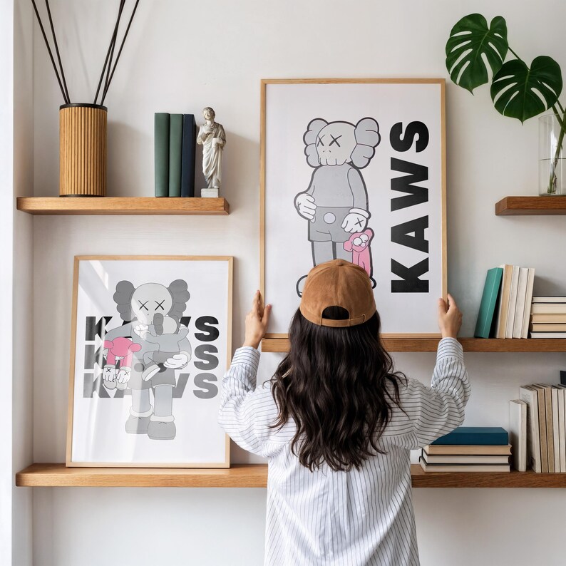 Puede incluir: Dos l&aacute;minas enmarcadas de arte KAWS con un personaje de dibujos animados en gris y rosa. La palabra "KAWS" est&aacute; impresa verticalmente en un marco. El otro marco tiene las letras "K", "A", "W" y "S" en blanco y negro. Las l&aacute;minas est&aacute;n en estantes de madera.