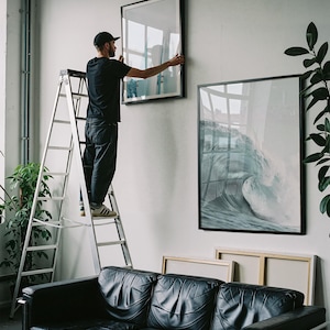 May include: A person on a ladder hangs a framed artwork on a white wall. The artwork features a wave crashing. A black leather sofa sits below the art. Additional framed art pieces are visible, along with potted plants.