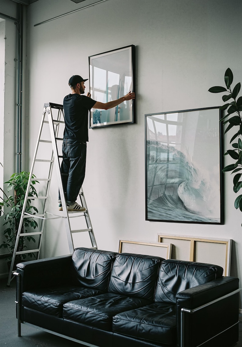 May include: A person on a ladder hangs a framed artwork on a white wall. The artwork features a wave crashing. A black leather sofa sits below the art. Additional framed art pieces are visible, along with potted plants.