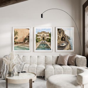 May include: A set of three framed wall art prints displayed above a beige modular sofa. The prints feature a Mediterranean pool terrace, a grand villa with a swimming pool, and a white vintage car driving along a coastal cliffside road.