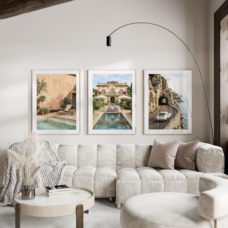 May include: A set of three framed wall art prints displayed above a beige modular sofa. The prints feature a Mediterranean pool terrace, a grand villa with a swimming pool, and a white vintage car driving along a coastal cliffside road.