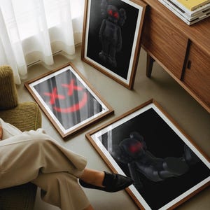 K&ouml;nnte beinhalten: Drei gerahmte Kunstdrucke mit von KAWS inspirierten Figuren mit roten 'XX'-Augen und einem roten Smiley. Die Drucke befinden sich in Holzrahmen und werden auf einem Boden und vor einem Holzschrank ausgestellt.