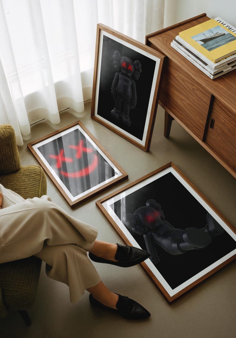 K&ouml;nnte beinhalten: Drei gerahmte Kunstdrucke mit von KAWS inspirierten Figuren mit roten 'XX'-Augen und einem roten Smiley. Die Drucke befinden sich in Holzrahmen und werden auf einem Boden und vor einem Holzschrank ausgestellt.