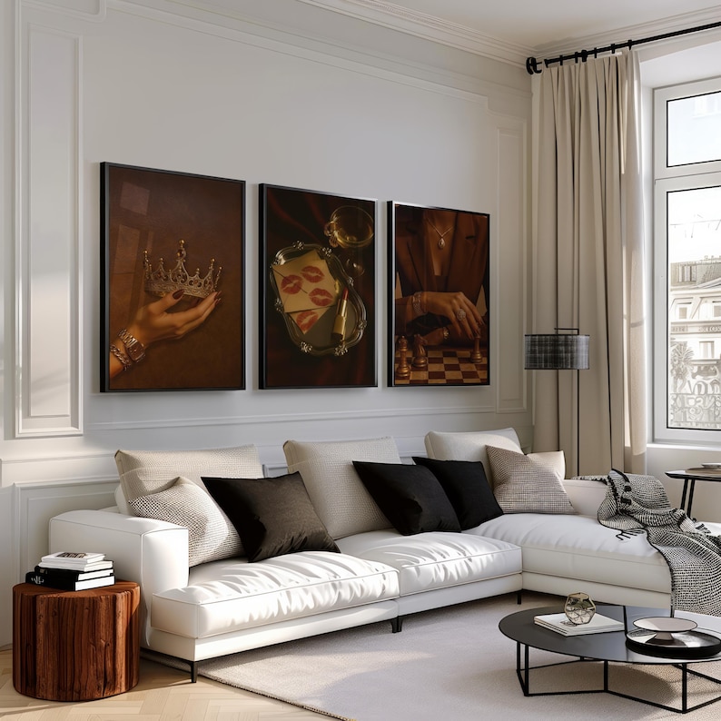 May include: Three framed art prints with a vintage aesthetic. One shows a hand holding a crown, another features lipstick kisses on a tray, and the third depicts a person playing chess. The prints are displayed above a white sectional sofa.
