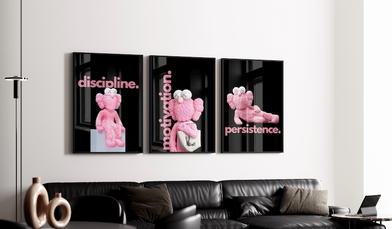 May include: Triptych of framed art prints featuring pink cartoon figures against a black background. Each print has a different word in pink: "discipline," "motivation," and "persistence." The prints are displayed above a black leather sofa.