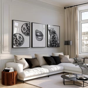 May include: Three framed black and white art prints. One features two pool balls with the number 8, another shows open lips, and the third depicts a leopard's back. The prints hang above a white sectional sofa.