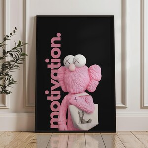 May include: A framed art print featuring a pink cartoon character with white eyes and a cross on its chest, holding a tote bag. The background is black with the word "motivation" in pink.