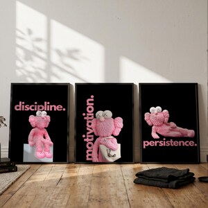 May include: Three framed art prints with black backgrounds and pink text. Each print features a pink cartoon character. The text reads "discipline.", "motivation.", and "persistence." The prints are displayed on a wooden floor.