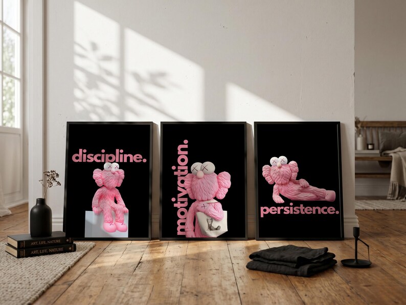 May include: Three framed art prints with black backgrounds and pink text. Each print features a pink cartoon character. The text reads "discipline.", "motivation.", and "persistence." The prints are displayed on a wooden floor.