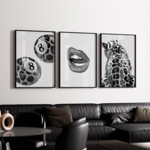 May include: Three framed monochrome art prints. The prints depict a leopard-print eight ball, a close-up of glossy lips, and the back of a giraffe. The prints are displayed above a black leather sofa in a living room.