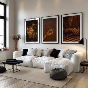 May include: Three framed art prints adorn a white sofa. The prints depict a lipstick kiss, a bejeweled crown, and a person playing chess. The room has a modern design with a black coffee table, a floor lamp, and a neutral rug.
