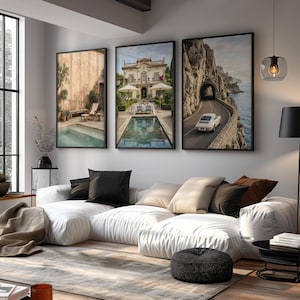 May include: A white modular sofa with black and beige throw pillows, positioned in front of three framed wall art prints. The prints depict a pool terrace, a villa with a pool, and a vintage car on a coastal road. A black floor lamp stands nearby.