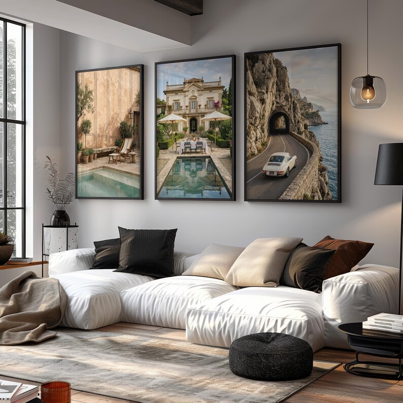 May include: A white modular sofa with black and beige throw pillows, positioned in front of three framed wall art prints. The prints depict a pool terrace, a villa with a pool, and a vintage car on a coastal road. A black floor lamp stands nearby.