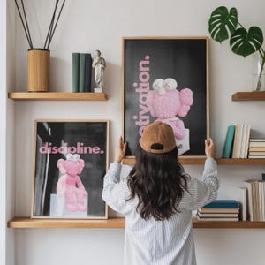 May include: Two framed art prints with a black background and pink text that reads "discipline" and "motivation." Each print features a pink cartoon character. The prints are displayed on wooden shelves.