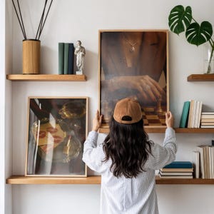 May include: A person holding a framed print of a chess game with a woman's hand. The print is displayed on a wooden shelf with other decorative items, including books, a statue, and a reed diffuser.