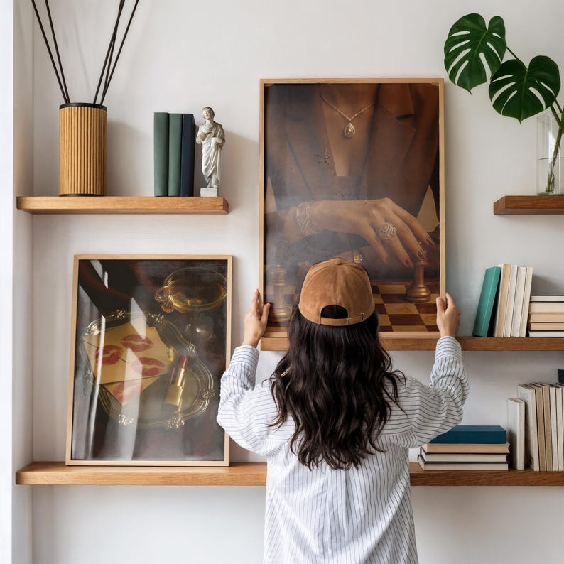 May include: A person holding a framed print of a chess game with a woman's hand. The print is displayed on a wooden shelf with other decorative items, including books, a statue, and a reed diffuser.