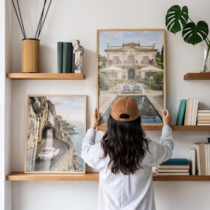 May include: A framed wall art print depicting a grand villa with a pool and patio, positioned on a wooden shelf next to a second framed print showing a white car driving on a coastal road. The scene includes books, a statue, and a plant.