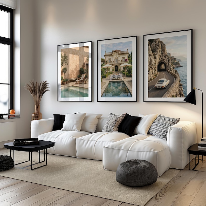 May include: Three framed wall art prints displayed above a white sectional sofa. The prints feature a sunlit pool terrace, a grand villa with a pool, and a vintage car driving along a coastal cliff road. The artwork is set in black frames.
