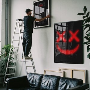 K&ouml;nnte beinhalten: Eine Person an einer Leiter h&auml;ngt gerahmte Kunst an einer wei&szlig;en Wand auf. Die Kunst umfasst ein Schwarz-Wei&szlig;-St&uuml;ck und ein schwarzes St&uuml;ck mit einem roten Smiley-Gesicht. Darunter befindet sich ein schwarzes Ledersofa.