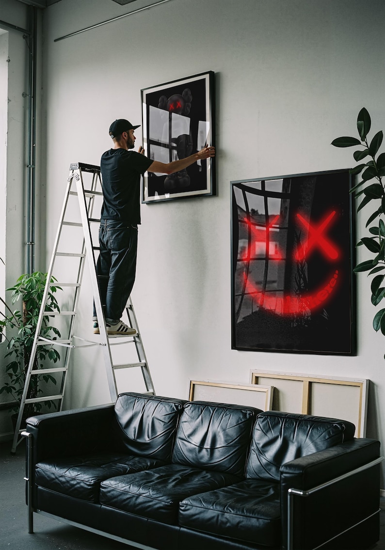 K&ouml;nnte beinhalten: Eine Person an einer Leiter h&auml;ngt gerahmte Kunst an einer wei&szlig;en Wand auf. Die Kunst umfasst ein Schwarz-Wei&szlig;-St&uuml;ck und ein schwarzes St&uuml;ck mit einem roten Smiley-Gesicht. Darunter befindet sich ein schwarzes Ledersofa.