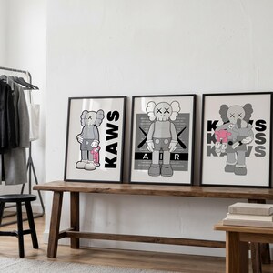 Puede incluir: Tres impresiones art&iacute;sticas enmarcadas con personajes KAWS en escala de grises. Las impresiones se exhiben en un banco de madera. El personaje KAWS se representa en varias poses, con la palabra "KAWS" o "AIR" y "KISS" en las impresiones.