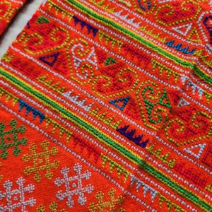 May include: A vibrant orange fabric featuring intricate, multi-colored cross-stitch embroidery. The textile displays horizontal bands of geometric patterns, including spirals, triangles, and cross shapes in shades of green, blue, yellow, and white.