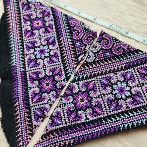 Puede incluir: Dos piezas de tela triangulares con intrincado bordado de punto de cruz en tonos morado, lavanda y negro. Una cinta métrica blanca está colocada a lo largo del borde superior, mostrando una longitud de aproximadamente 23 cm para la tela.