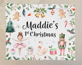 Könnte beinhalten: Eine weiße Weihnachtsdecke mit dem Text "Maddie's 1st Christmas". Die Decke zeigt Illustrationen einer Ballerina, eines Schwans, eines Nussknackers und Weihnachtsschmuck. Das Design umfasst einen Weihnachtsbaum, einen Lebkuchenmann und Stechpalmen.
