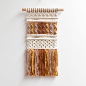 May include: A macrame wall hanging with a wooden dowel. The woven piece features cream, pink, orange, and brown colors. The design includes spiral and fringe details, creating a textured, bohemian aesthetic. This handcrafted wall decor adds a touch of warmth and style.