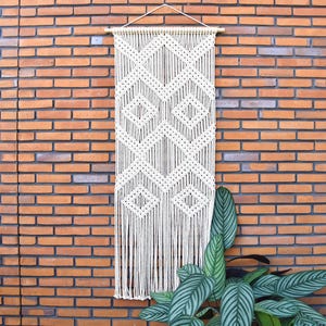 May include: A cream-colored macrame wall hanging with a geometric diamond pattern. The wall decor is suspended from a wooden dowel and features long fringe at the bottom. The background is a red brick wall, with green leafy plants in the lower right corner.