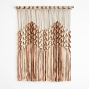 May include: A macrame wall hanging featuring a geometric design in shades of cream, tan, and brown. The piece is suspended from a wooden dowel and has long fringe at the bottom, adding texture and visual interest to the decor.