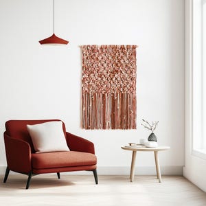 May include: A modern living room scene featuring a rust-colored armchair with a white pillow, a small wooden side table, and a decorative wall hanging with a textured, fringed design in shades of red and cream.