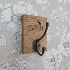 May include: A wooden coat hook with a single black metal hook. The wood is stained a dark brown and has the name "PADDY" engraved on it, along with a small dinosaur silhouette.