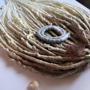 Puede incluir: Conjunto de rastas hechas a mano en tonos beige y marrón, dispuestas sobre una superficie blanca. Un pequeño accesorio para el cabello enrollado en azul y blanco, y una pequeña piedra también están presentes. Una cinta marrón añade un toque decorativo.
