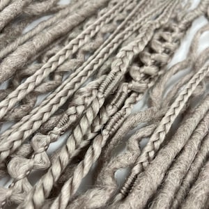 Puede incluir: Primer plano de múltiples rastas de color marrón claro, algunas con secciones trenzadas y nudos decorativos. Las rastas varían en grosor y textura, creando una apariencia natural y hecha a mano. El fondo es una superficie blanca lisa, que resalta los detalles de las extensiones de cabello.