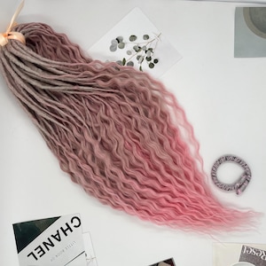 May include: A bundle of wavy, two-toned synthetic dreadlocks. The top portion is a light brown, transitioning to a pink hue that deepens towards the ends. A small, beige ribbon secures the top. A hair tie is also visible.