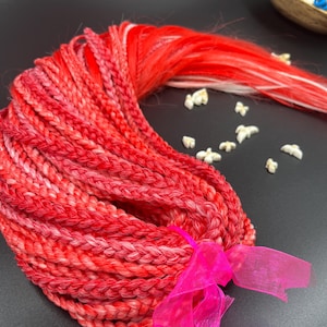 Könnte beinhalten: Ein Bündel aus rot- und rosafarbenen geflochtenen Haarverlängerungen, gebunden mit einem leuchtend rosa Band. Das Haar hat eine Mischung aus roten und rosa Farbtönen, mit einigen weißen Strähnen. Der Hintergrund ist schwarz.