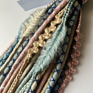 Puede incluir: Un paquete de rastas sintéticas en varios tonos de azul, beige y rosa. Las rastas están atadas juntas con una cinta azul y una banda elástica negra.