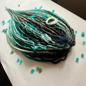 Puede incluir: Una colección de rastas de color turquesa, marrón y negro con detalles en blanco y azul. Las rastas están adornadas con una goma para el pelo blanca, tela azul y pequeñas cuentas decorativas azules y doradas. Dispuestas sobre una superficie blanca.