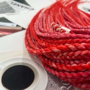 May include: A close-up of a bundle of red and pink synthetic hair extensions. The extensions are braided and have a white string running through them.