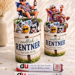 May include: Two cylindrical containers featuring illustrations of a man and a woman relaxing in lounge chairs with drinks. The containers are labeled "endlich RENTNER - Ich habe fertig" and include two rolled paper scrolls with German text and graphics.