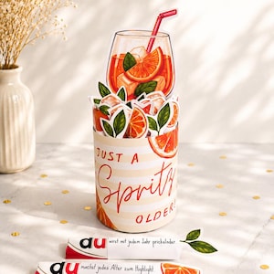 Peut inclure: Une carte de vœux cylindrique présentant une illustration en relief d'un cocktail spritz orange avec une paille, des glaçons et des tranches d'agrumes. La carte porte l'inscription "JUST A Spritz OLDER!" et deux petits inserts en papier.