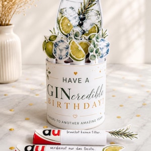 May include: A cylindrical birthday card featuring a pop-up illustration of a gin and tonic with lime and ice. The card reads "HAVE A GINcredible BIRTHDAY! CHEERS TO ANOTHER AMAZING YEAR!" Two small wrapped items with German text are placed in front.