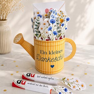 Puede incluir: Una regadera amarilla con rayas blancas que contiene recortes de papel de flores silvestres y un sobre blanco. La regadera tiene el texto "Ein kleines Dankeschön" y un corazón azul. Dos rollos de papel con extremos florales yacen delante.