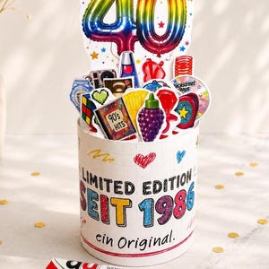 Könnte beinhalten: Ein zylindrischer Behälter mit der Aufschrift LIMITED EDITION SEIT 1986 ein Original, gefüllt mit bunten Ausschnitten wie einem großen Regenbogen-Ballon 40, einem Zauberwürfel, einer traubenförmigen Figur und einem 90's HITS Schild, sowie einem Papierstreifen mit der Aufschrift warst dabei, als Handys Antennen hatten.