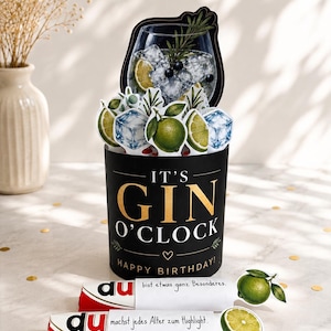 May include: A black cylindrical greeting card featuring the gold text IT'S GIN O'CLOCK and HAPPY BIRTHDAY. The top displays a pop-up illustration of a gin cocktail with ice, lime slices, and rosemary. Two white paper strips with German text are attached.