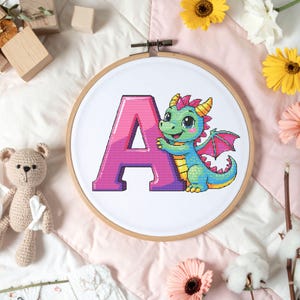 Könnte beinhalten: Ein Kreuzstich-Stickrahmen mit dem rosa Buchstaben „A“ und einem Zeichentrickdrachen. Der Drache ist grün und blau mit rosa Flügeln und einem gelb-rosa Kamm. Der Rahmen befindet sich vor einem weißen Hintergrund.