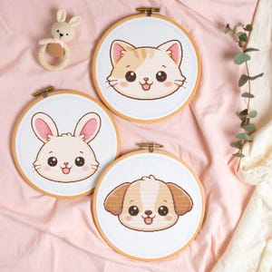 Könnte beinhalten: Drei bestickte Reifen mit niedlichen Tiergesichtern: eine Katze, ein Hase und ein Welpe. Die Katze ist hellbraun und weiß, der Hase ist weiß mit rosa Ohren und der Welpe ist hellbraun und weiß. Ein kleiner Häkel-Hasen-Rassel ist ebenfalls im Bild.